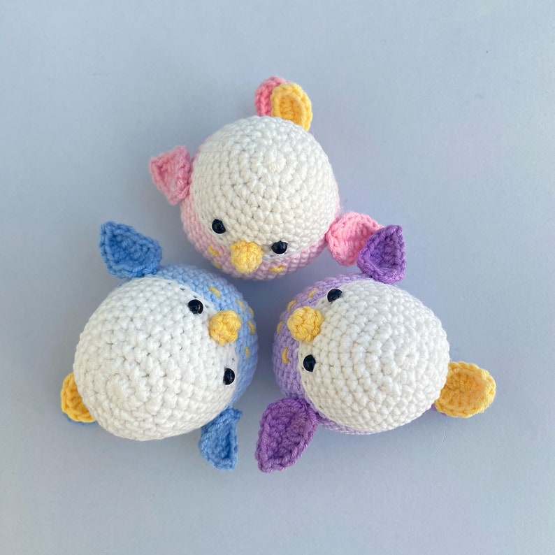 May include: Three handmade crochet bird toys in white, pink, blue, and purple. Each bird has a white body, black eyes, and a yellow beak. The wings are in coordinating colors. These soft toys are perfect for children.