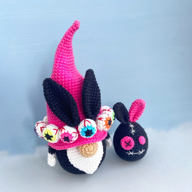 K&ouml;nnte beinhalten: Ein rosa und schwarzer H&auml;kel-Zwerg mit einem Hut, der mit bunten Augen verziert ist. Neben dem Zwerg befindet sich ein kleiner schwarzer H&auml;kel-Hase mit einem gestickten Gesicht.