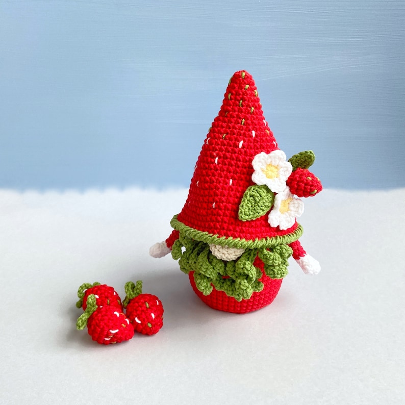May include: A crocheted strawberry gnome with a green beard and white flowers. Three smaller crocheted strawberries are in the foreground.
