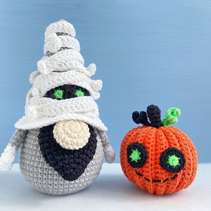 Pu&ograve; includere: Un gnomo mummia all'uncinetto con un cappello bianco all'uncinetto e una barba nera, accanto a una zucca arancione all'uncinetto con occhi verdi e uno stelo nero.