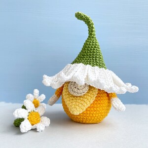 May include: A crocheted gnome with a green hat and white beard, holding a yellow and white crocheted flower. The gnome is sitting on a white surface.