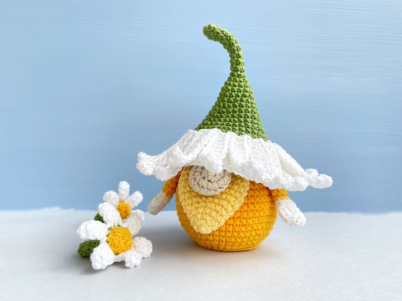 May include: A crocheted gnome with a green hat and white beard, holding a yellow and white crocheted flower. The gnome is sitting on a white surface.