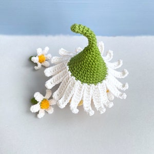 May include: A crocheted daisy flower with a green center and white petals. The flower is topped with a green crocheted hat. There are two smaller crocheted daisies on the side.