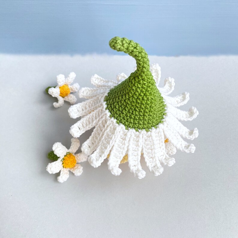 May include: A crocheted daisy flower with a green center and white petals. The flower is topped with a green crocheted hat. There are two smaller crocheted daisies on the side.