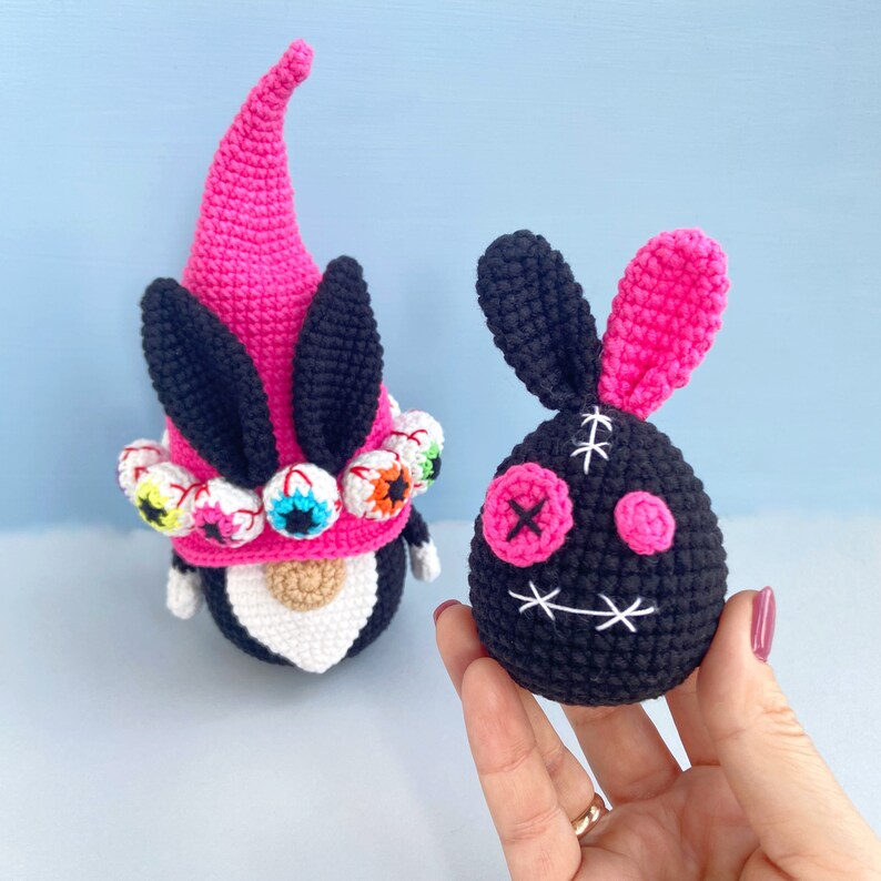 K&ouml;nnte beinhalten: Zwei geh&auml;kelte Halloween-Spielzeuge. Eines ist ein rosa-schwarzer Zwerg mit wei&szlig;em Bart und bunten Augen. Das andere ist ein rosa-schwarzes Kaninchen mit einem gestickten Gesicht und einem gro&szlig;en rosa Auge.