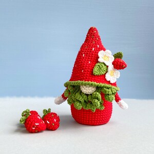 May include: A crocheted strawberry gnome with a green beard and white flowers. The gnome is standing on a white surface with two smaller crocheted strawberries.
