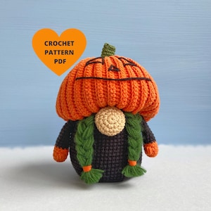 Può includere: Schema a uncinetto PDF per un gnomo zucca con corpo nero, trecce verdi e un cappello di zucca arancione.