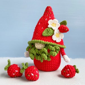 May include: A crocheted strawberry gnome with a green beard and white flowers on its hat. Three smaller crocheted strawberries are in the foreground.