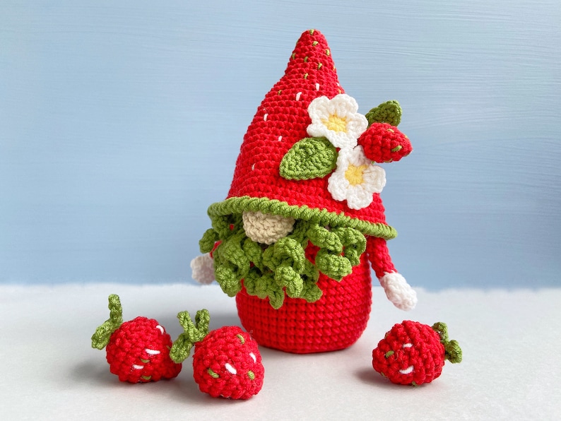 May include: A crocheted strawberry gnome with a green beard and white flowers on its hat. Three smaller crocheted strawberries are in the foreground.