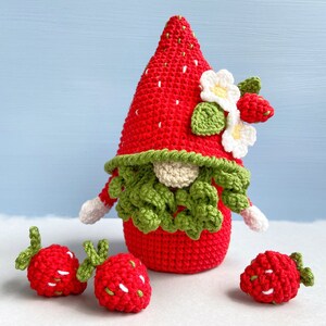 May include: A crocheted strawberry gnome with a red and white hat and green beard. The gnome is surrounded by three smaller crocheted strawberries.