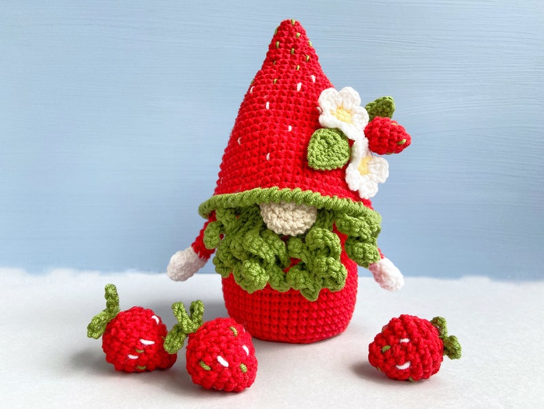 May include: A crocheted strawberry gnome with a red and white hat and green beard. The gnome is surrounded by three smaller crocheted strawberries.