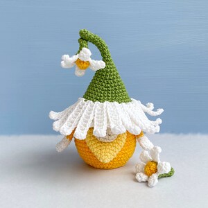May include: A crocheted gnome with a green hat and white trim. The gnome is holding a yellow flower with white petals.