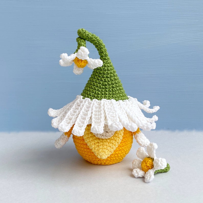 May include: A crocheted gnome with a green hat and white trim. The gnome is holding a yellow flower with white petals.