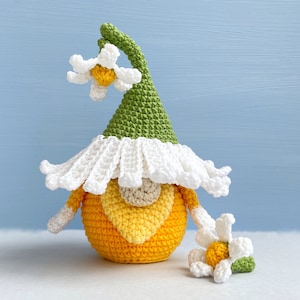 May include: A crocheted gnome with a yellow and orange body, a white hat with green accents, and a white flower on top. The gnome is holding a smaller white flower in its hand.