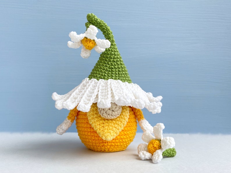 May include: A crocheted gnome with a yellow and orange body, a white hat with green accents, and a white flower on top. The gnome is holding a smaller white flower in its hand.