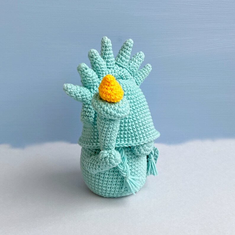 May include: A crocheted statue of the Statue of Liberty, holding a torch, in a light blue color.