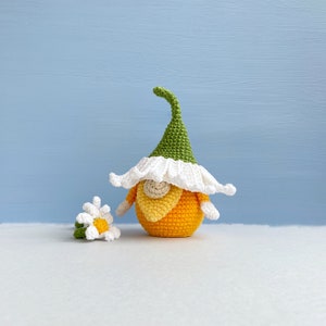 May include: A crocheted gnome with a green hat, white beard, and orange body. The gnome is holding a small crocheted white flower with a yellow center.