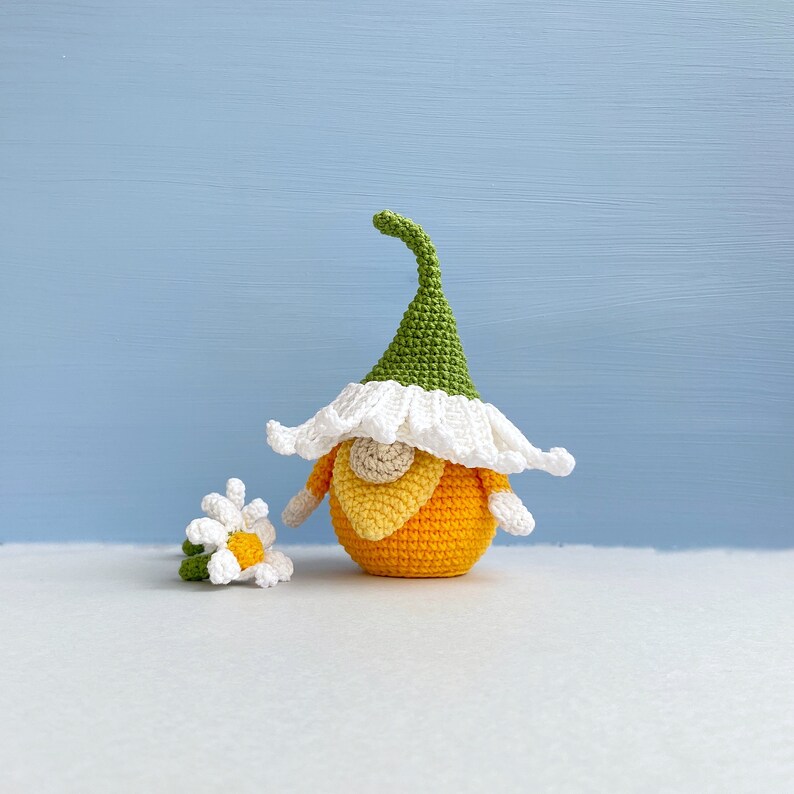 May include: A crocheted gnome with a green hat, white beard, and orange body. The gnome is holding a small crocheted white flower with a yellow center.