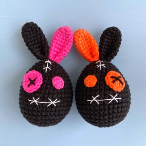 May include: Two crocheted bunny plushies with black bodies, pink and orange ears, and stitched-on eyes and mouths.