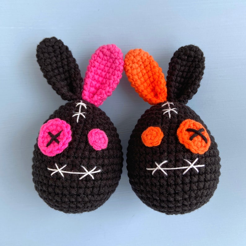 May include: Two crocheted bunny plushies with black bodies, pink and orange ears, and stitched-on eyes and mouths.