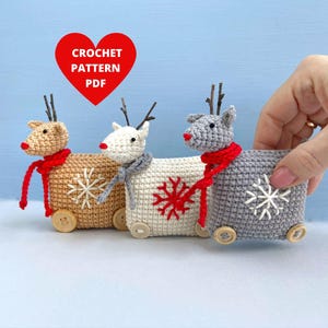 May include: Three crocheted reindeer toys on wheels. The reindeer are brown, white, and gray with red scarves and snowflake designs. The toys are on a light blue background.