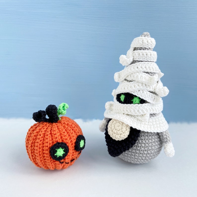 Pu&ograve; includere: Un gnomo di Halloween all'uncinetto che indossa un costume da mummia bianco e una zucca arancione all'uncinetto con occhi verdi.