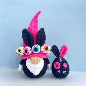 K&ouml;nnte beinhalten: Ein rosa-schwarzer H&auml;kel-Zwerg mit wei&szlig;em Bart und einer M&uuml;tze, die mit bunten Augen verziert ist. Neben dem Zwerg befindet sich ein kleiner schwarzer H&auml;kel-Hase mit einem aufgestickten Gesicht.