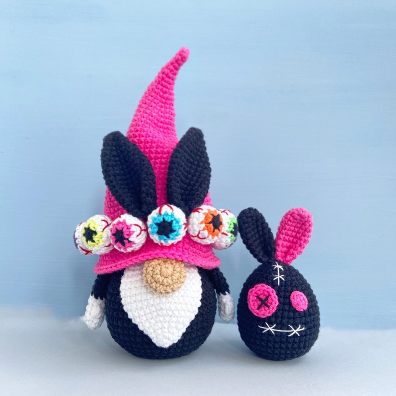 K&ouml;nnte beinhalten: Ein rosa-schwarzer H&auml;kel-Zwerg mit wei&szlig;em Bart und einer M&uuml;tze, die mit bunten Augen verziert ist. Neben dem Zwerg befindet sich ein kleiner schwarzer H&auml;kel-Hase mit einem aufgestickten Gesicht.