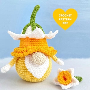 May include: A crocheted daffodil gnome with a white and yellow hat and a white beard. The gnome is holding a smaller crocheted daffodil flower. The text "CROCHET PATTERN PDF" is in a yellow heart shape.