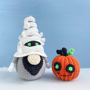 Pu&ograve; includere: Un gnomo mummia all'uncinetto con un cappello di bendaggio bianco e una barba nera si trova accanto a una zucca arancione all'uncinetto con occhi verdi e uno stelo.
