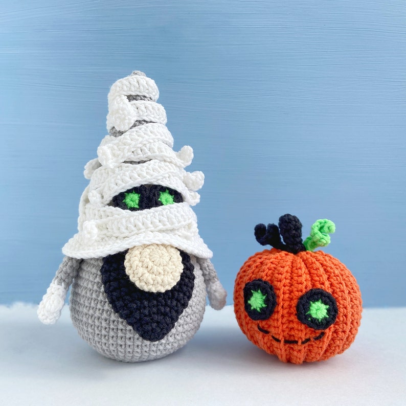 Pu&ograve; includere: Un gnomo mummia all'uncinetto con un cappello di bendaggio bianco e una barba nera si trova accanto a una zucca arancione all'uncinetto con occhi verdi e uno stelo.