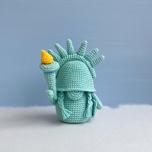 May include: A crocheted Statue of Liberty figurine with a teal green body and a yellow torch. The figurine is standing on a white surface.
