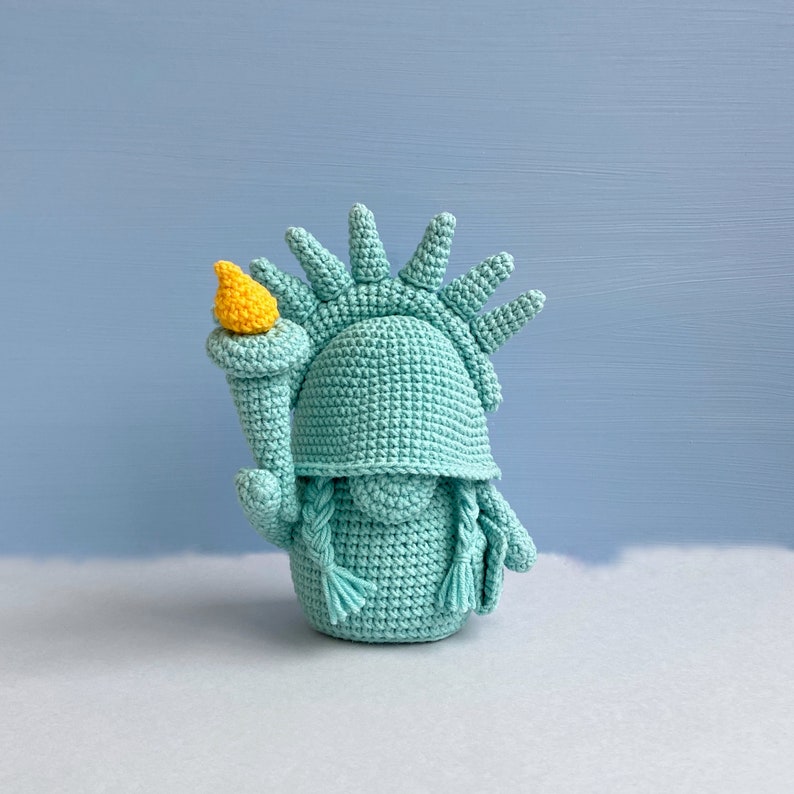 May include: A crocheted Statue of Liberty figurine with a teal green body and a yellow torch. The figurine is standing on a white surface.