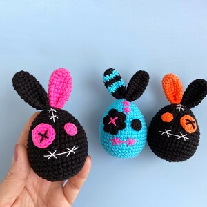 May include: Three crocheted Easter bunny ornaments. One is black with pink accents, one is blue with white accents, and one is black with orange accents. Each bunny has a stitched-on face with X-shaped eyes.