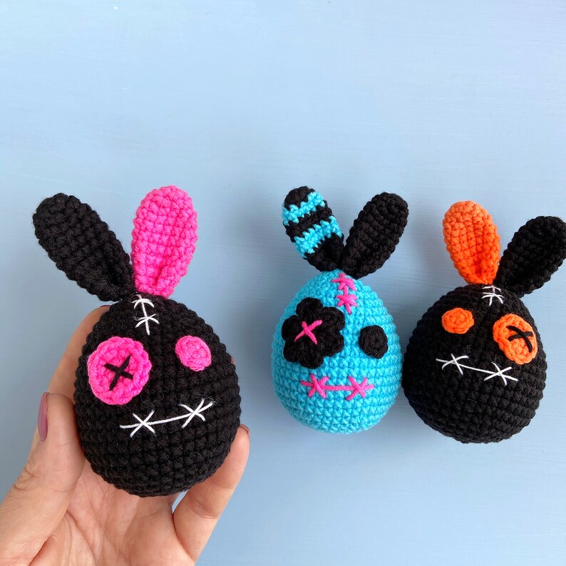 May include: Three crocheted Easter bunny ornaments. One is black with pink accents, one is blue with white accents, and one is black with orange accents. Each bunny has a stitched-on face with X-shaped eyes.