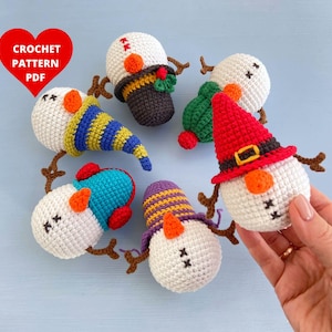 Pode incluir: Seis enfeites de boneco de neve de crochê com chapéus e cachecóis de diferentes cores. Os enfeites estão dispostos em círculo sobre um fundo azul claro. O texto "CROCHET PATTERN PDF" está em um coração vermelho.