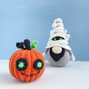 Pu&ograve; includere: Una zucca arancione all'uncinetto con foglie verdi e occhi neri, accanto a un gnomo mummia bianco all'uncinetto con la barba nera.