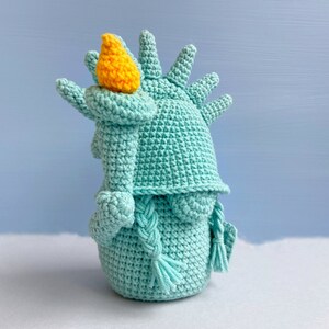 May include: A crocheted plush toy shaped like the Statue of Liberty. The toy is teal blue with a yellow torch and a braided rope for hair.
