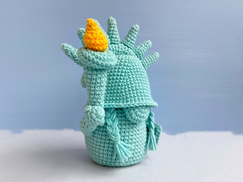 May include: A crocheted plush toy shaped like the Statue of Liberty. The toy is teal blue with a yellow torch and a braided rope for hair.
