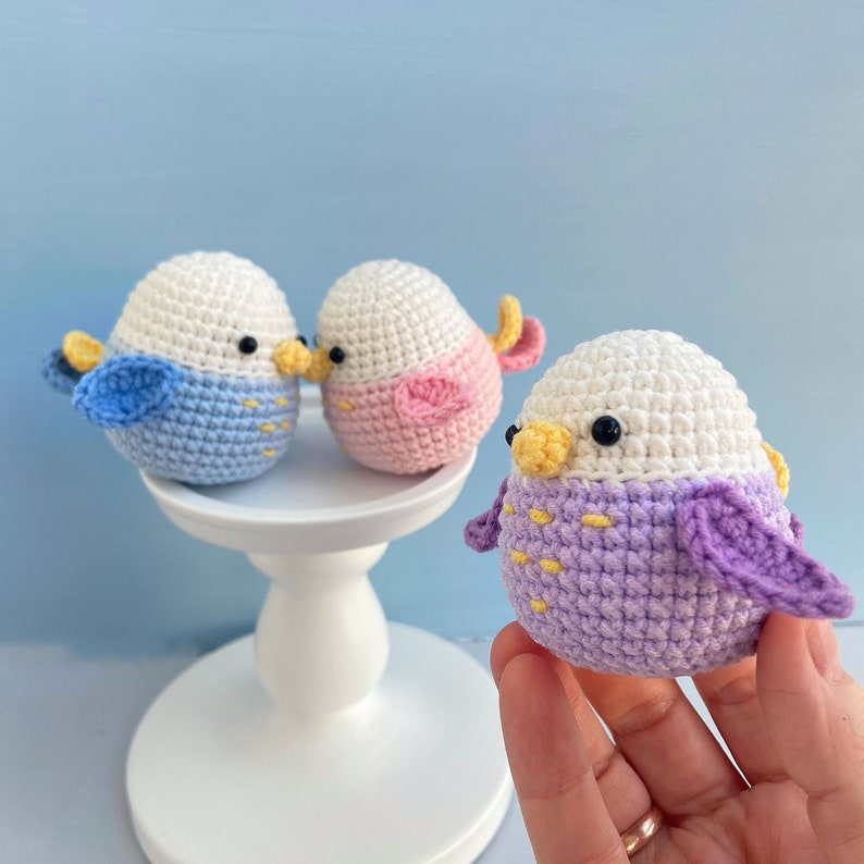 May include: Crocheted bird ornaments in blue, pink, and purple with white heads, black eyes, and yellow beaks. The birds have small wings and are displayed on a white pedestal and held by a hand. The background is a light blue.