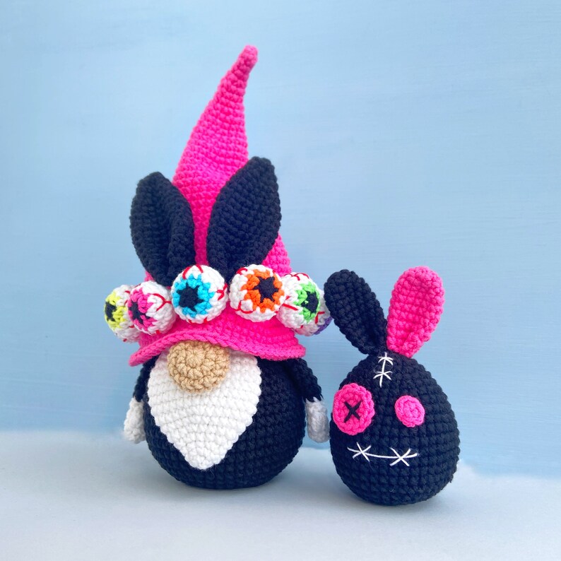 K&ouml;nnte beinhalten: Zwei geh&auml;kelte Gnome, einer gro&szlig; und einer klein. Der gr&ouml;&szlig;ere Gnome hat einen rosa Hut mit bunten Augen und einen schwarzen K&ouml;rper. Der kleinere Gnome ist schwarz mit rosa Ohren und einem gestickten Gesicht.