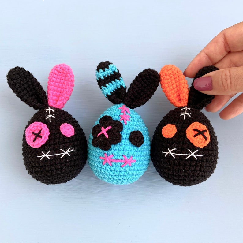 May include: Three crocheted bunny-shaped Easter eggs with stitched-on facial features. One egg is black with pink accents, one is blue with black and pink accents, and one is black with orange accents.