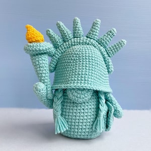 May include: A crocheted plush toy shaped like the Statue of Liberty. The toy is green with a yellow torch and a crown of spikes.