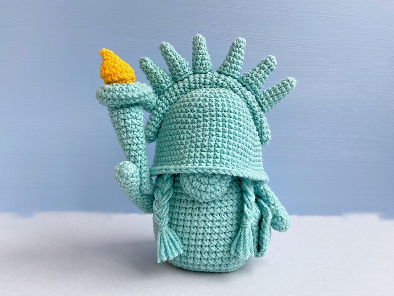May include: A crocheted plush toy shaped like the Statue of Liberty. The toy is green with a yellow torch and a crown of spikes.