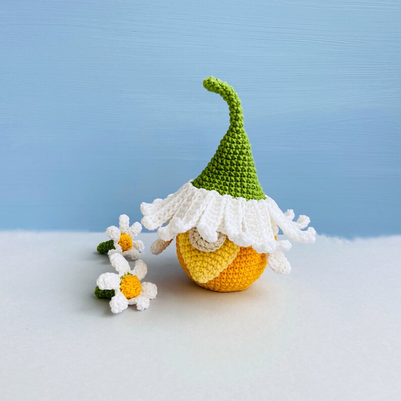 May include: A crocheted gnome with a green hat and white trim, sitting on a white surface with two crocheted daisies.