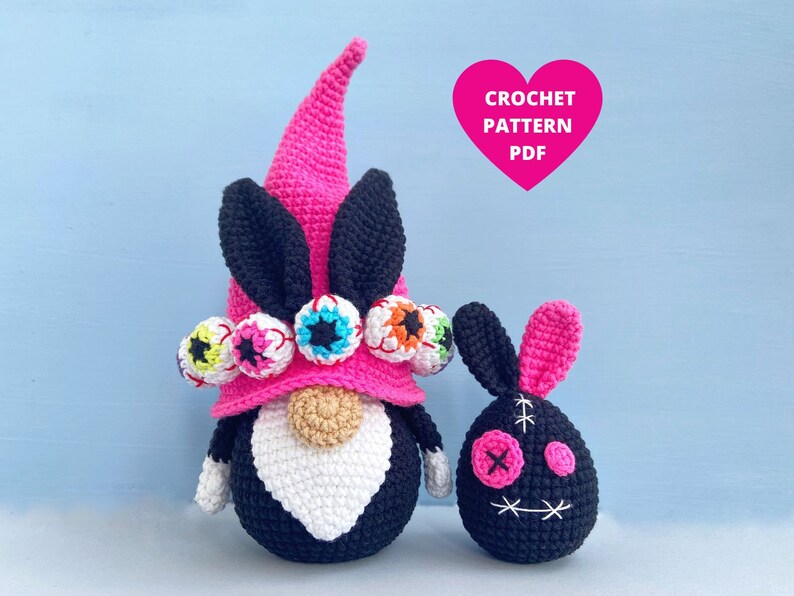 K&ouml;nnte beinhalten: Ein rosa und schwarzer geh&auml;kelter Zwerg mit einem Hut, der mit bunten Augen verziert ist. Neben dem Zwerg befindet sich ein kleinerer rosa und schwarzer geh&auml;kelter Hase mit einem gestickten Gesicht. Der Text "CROCHET PATTERN PDF" befindet sich in einem rosa Herz.