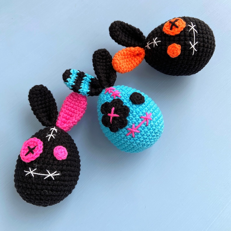 May include: Three crocheted Easter bunny eggs with stitched-on eyes and mouths. The eggs are black, blue, and pink with black and white stitching.