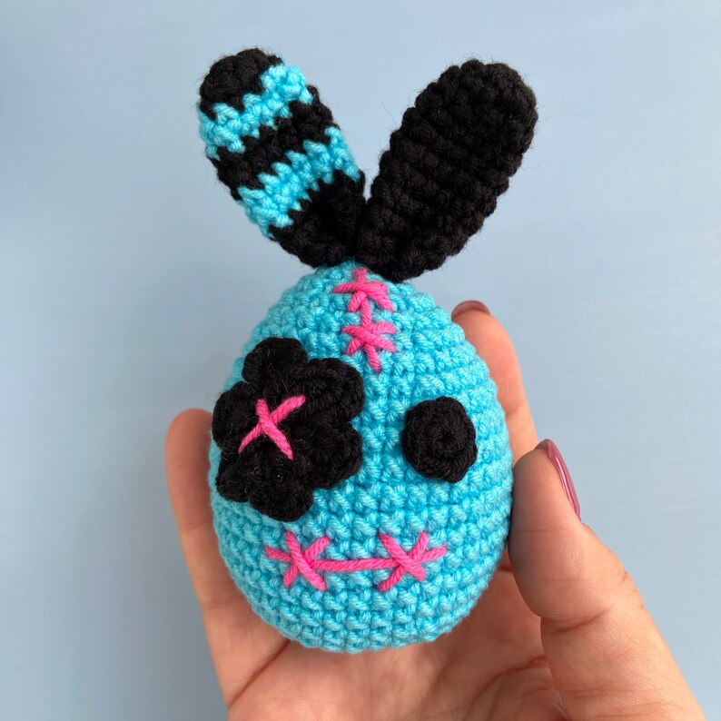 May include: A blue crocheted bunny with black ears and a stitched-on face. The bunny has pink stitches for a mouth and black felt eyes.