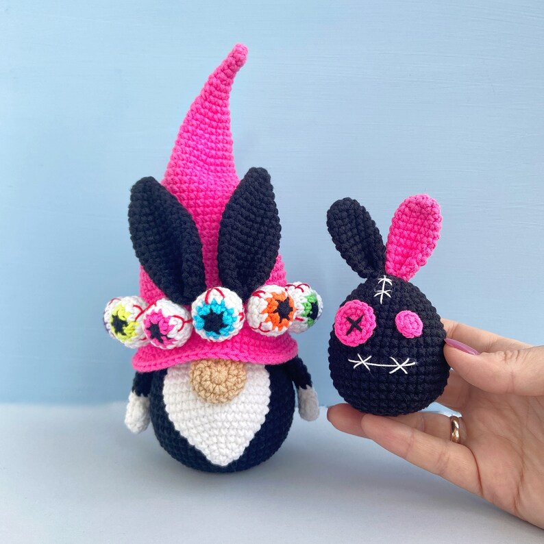 K&ouml;nnte beinhalten: Ein rosa-schwarzer H&auml;kel-Zwerg mit einem rosa Hut und schwarzen Ohren. Der Hut ist mit bunten Augen verziert. Ein schwarzer H&auml;kel-Hase mit rosa Ohren und einem gestickten Gesicht wird in einer Hand gehalten.