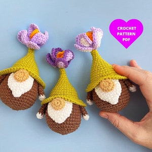 May include: Three crocheted gnomes with purple and white flowers on their hats. The gnomes are brown and have white beards. The background is a light blue.